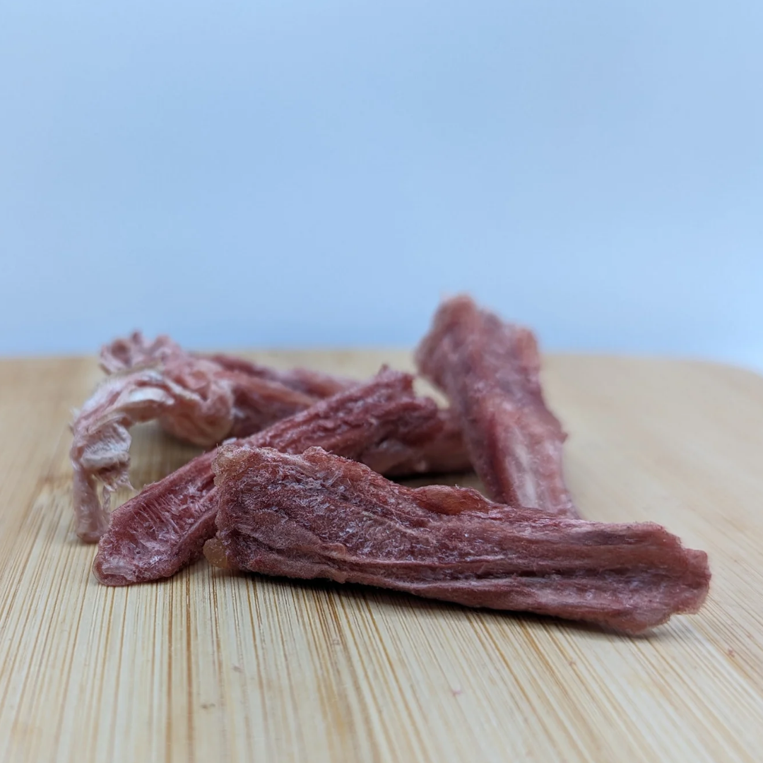 Four pieces of Freeze-dried Duck Tongues 1oz by Wild Nosh Pets, resembling a protein-packed treat, are placed on a wooden surface. The meat appears to be fibrous and textured, with a reddish-brown color.