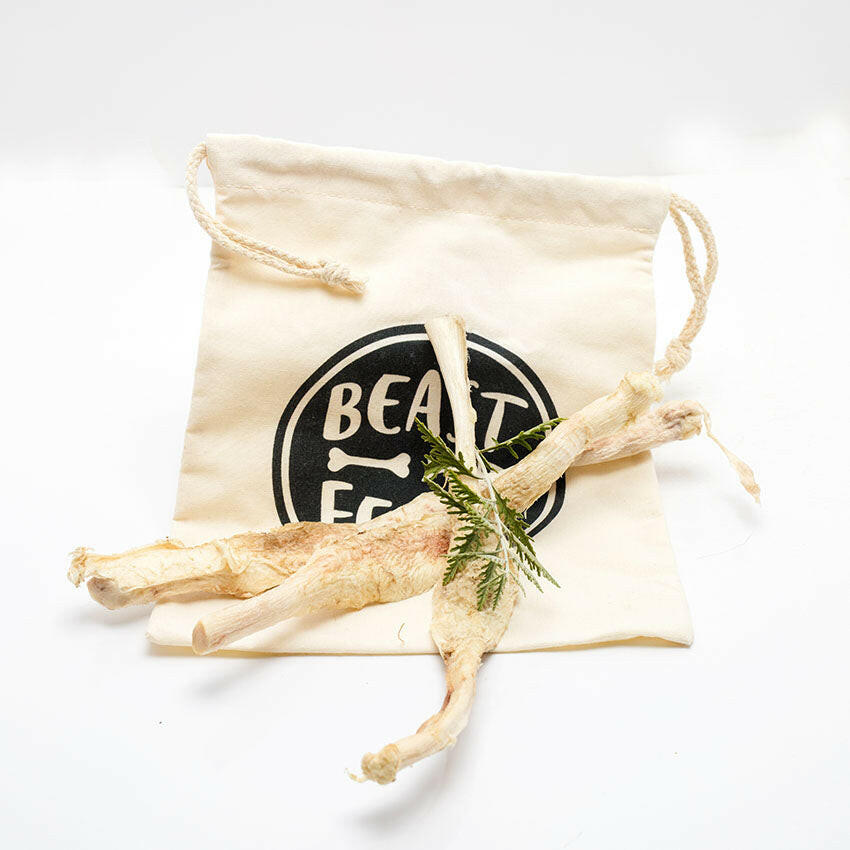 Freeze-dried bison patellar tendons from Wild Nosh Pets, ideal for teething puppies, rest on a beige drawstring bag featuring the "BEAST" label and bone logo. A sprig of greenery completes the natural touch.