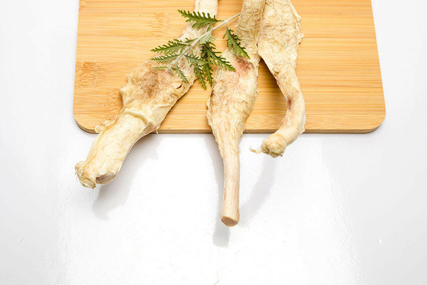 Dried licorice roots and green leaves rest on a wooden board atop a white surface, while teething puppies contentedly chew on Freeze-Dried Bison Patellar Tendons by Wild Nosh Pets.