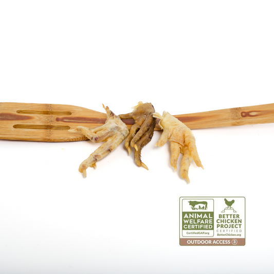 Three Wild Nosh Pets Dehydrated Chicken Feet on a wooden spatula, with Certified Humane and Better Chicken Commitment certification labels visible in the foreground.