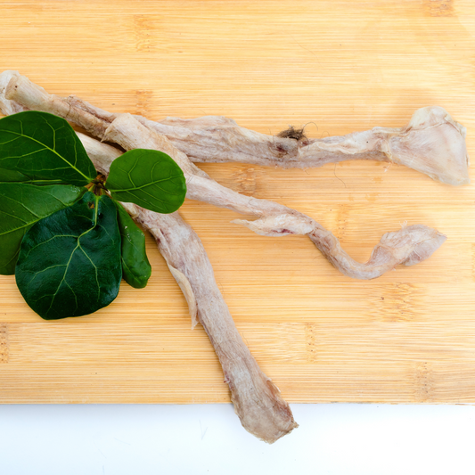 Two Wild Nosh Pets Freeze-Dried Bison Tibial Tendons, ideal for teething puppies who need extra collagen, rest on a light wooden cutting board beside three fresh green leaves.