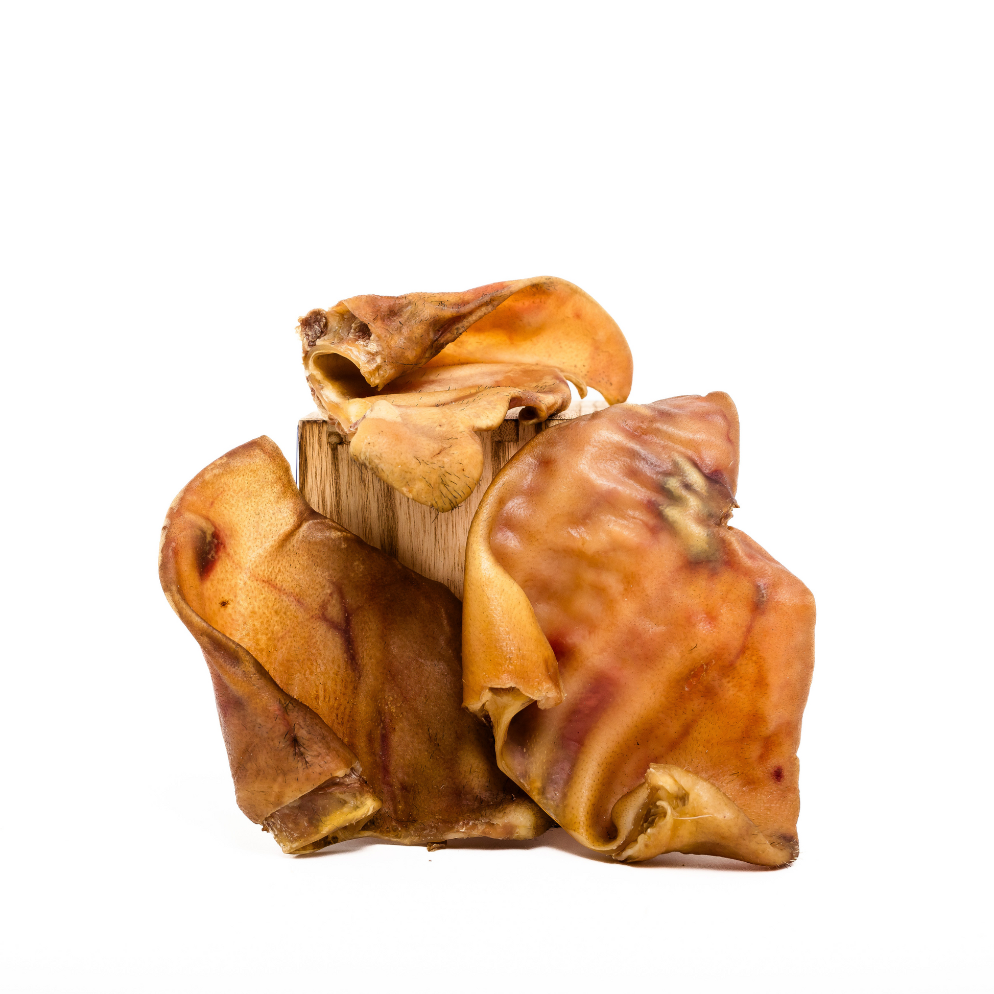 Three Wild Nosh Pets Heritage Breed Dehydrated Pig Ears, rich in glucosamine and chondroitin, are stacked on a wooden block against a white background.