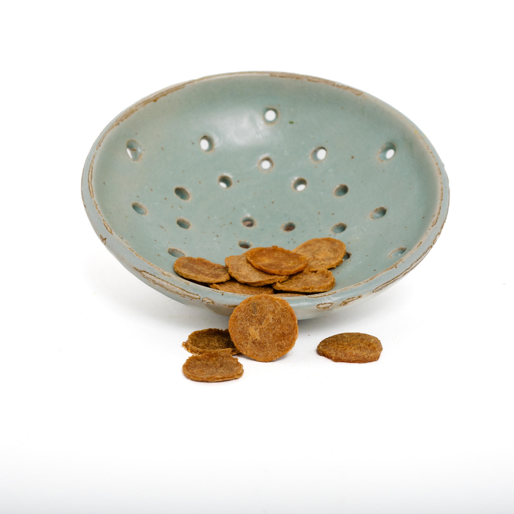 A light blue ceramic bowl with circular holes holds Wild Nosh Pets Carp Crispers 1 oz.—high-protein, omega-rich treats—some scattered outside the bowl, set against a white background.