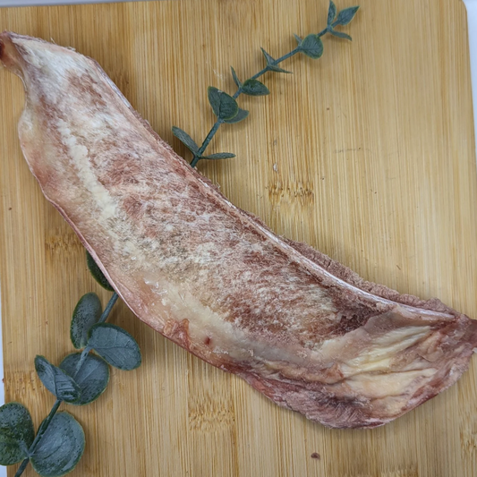 A slab of raw meat, possibly beef or Wild Nosh Pets Freeze-Dried Bison Scapula Slice, placed on a wooden cutting board with a sprig of herbs beside it.