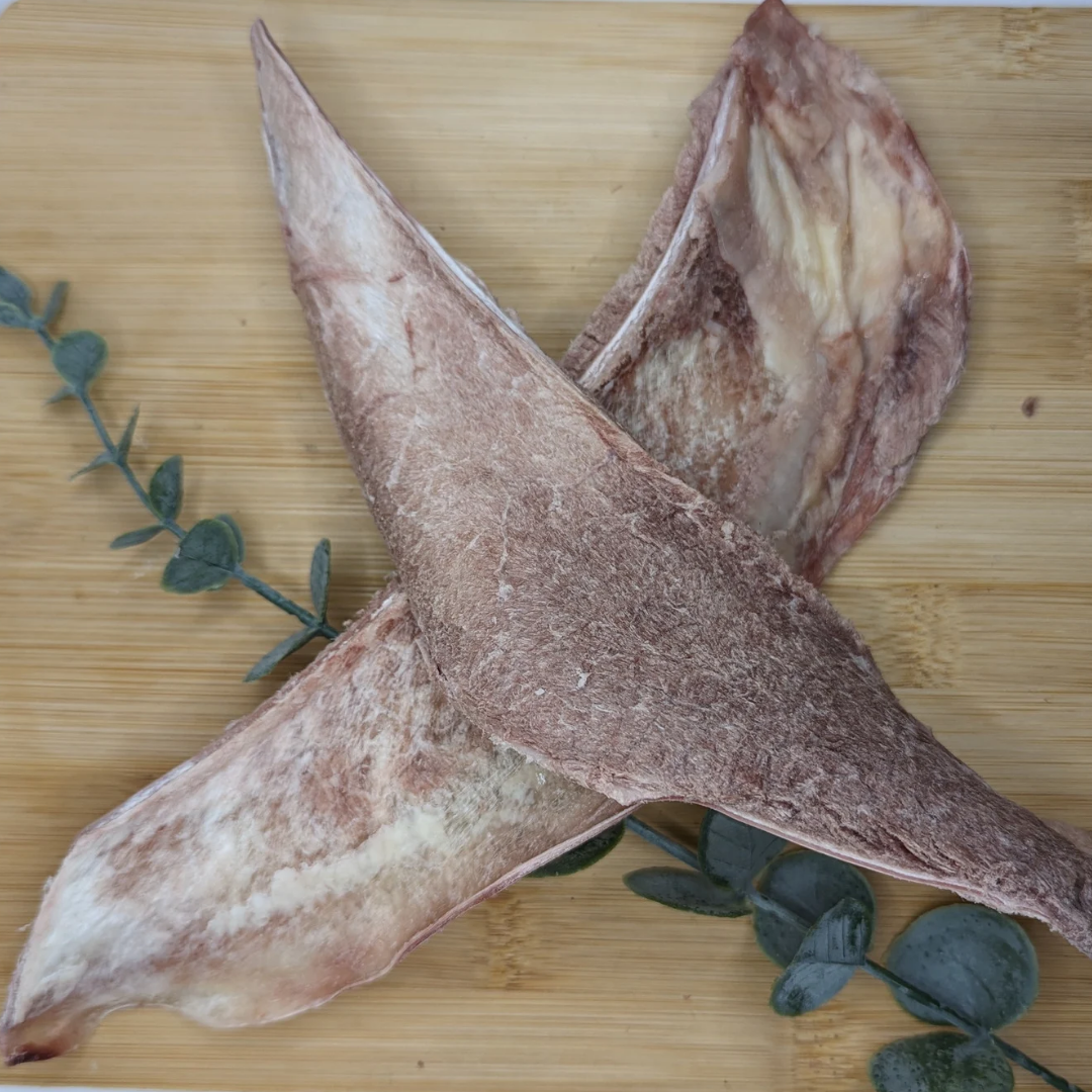 Two Freeze-Dried Bison Scapula Slices by Wild Nosh Pets are placed on a wooden cutting board next to a few sprigs of green herbs, perfect for senior dogs.