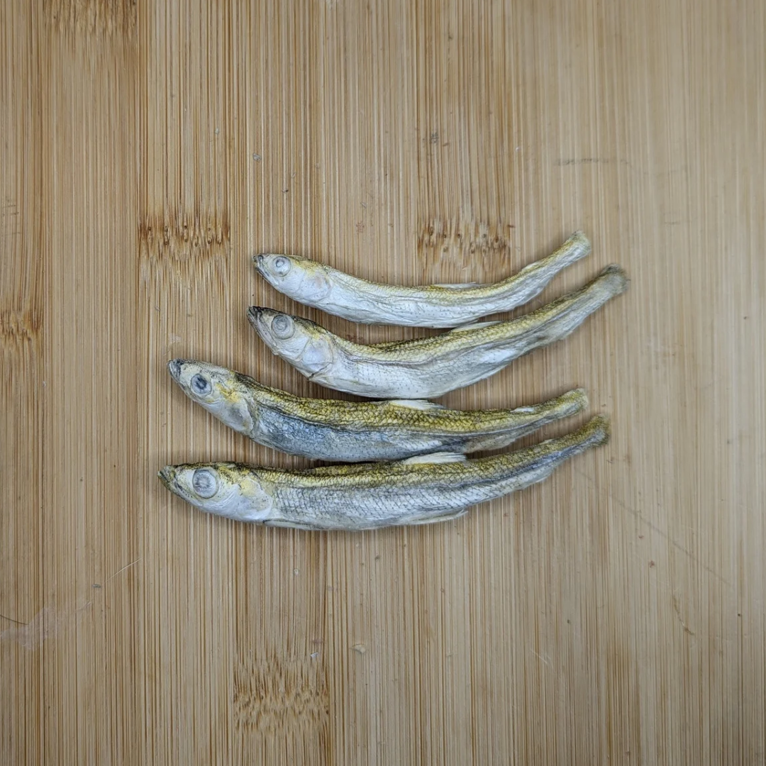 Four small, **Wild Nosh Pets Freeze-dried Smelt 1oz** laid out in a line on a wooden cutting board, showcasing a sustainable seafood option rich in Omega-3 fatty acids.