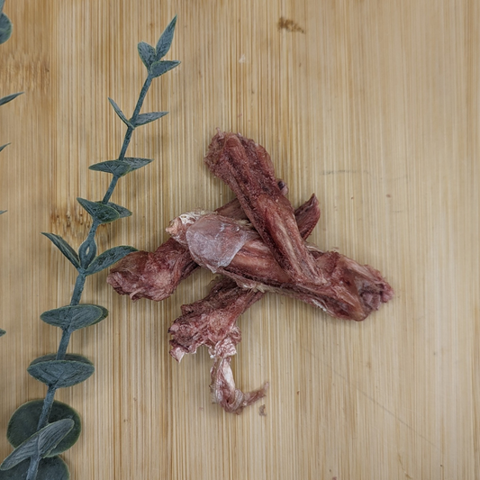 A small pile of Wild Nosh Pets Freeze-dried Duck Tongues 1oz next to a green leafy plant on a wooden surface.