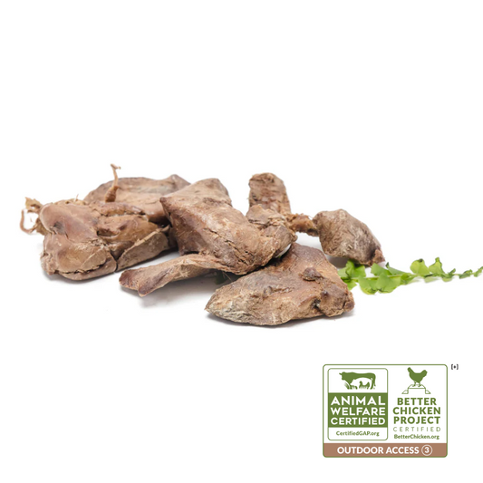 Several pieces of Freeze-Dried Chicken Liver 3oz by Wild Nosh Pets are scattered with a small green plant on a white surface. There are two certification labels in the bottom-right corner indicating Certified Humane Raised and Handled and Better Chicken Commitment standards.