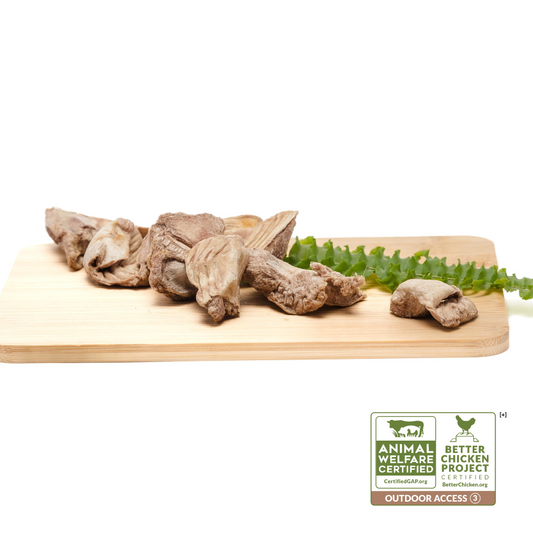 A wooden cutting board displaying pieces of meat, including Freeze-Dried Chicken Gizzards 3oz from Wild Nosh Pets, and a green vegetable. Certification logos for Animal Welfare and Better Chicken Project with outdoor access are present in the bottom right corner.