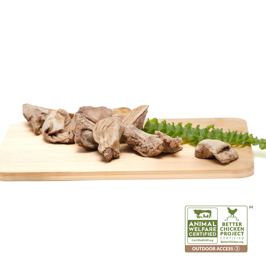 A wooden cutting board displaying pieces of meat, including Freeze-Dried Chicken Gizzards 3oz from Wild Nosh Pets, and a green vegetable. Certification logos for Animal Welfare and Better Chicken Project with outdoor access are present in the bottom right corner.