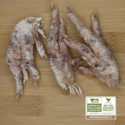 Three raw Wild Nosh Pets Freeze-Dried Chicken Feet on a wooden surface, accompanied by certification labels: Animal Welfare Certified and Better Chicken Commitment.