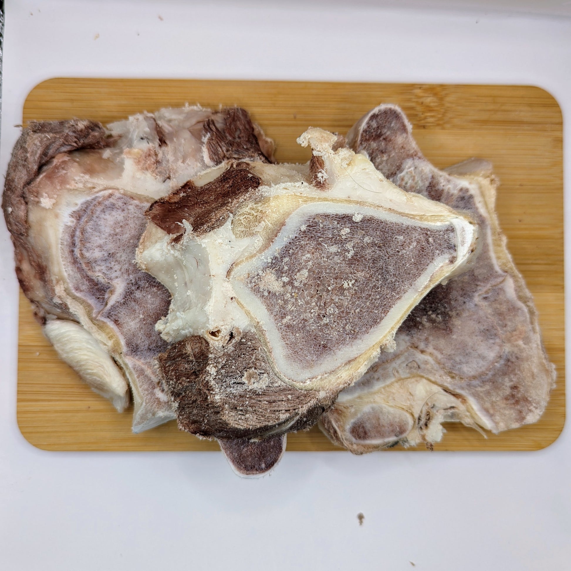 Frozen pork chops stacked on a wooden cutting board, alongside some tempting Wild Nosh Pets Freeze-Dried Bison Knuckle Slices.