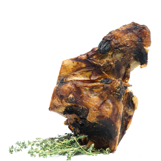 A large, cooked Smoked Bison Knuckle Bone from Wild Nosh Pets, browned and crispy, stands upright on a garnish of fresh thyme sprigs against a white background, showcasing its elegance.