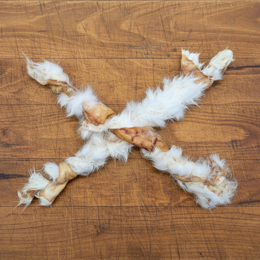 Two Wild Nosh Pets Rabbit Twists, rawhide dog chews with fur made from novel protein rabbit, are arranged in an X shape on a wooden surface—perfect as natural dog treats.